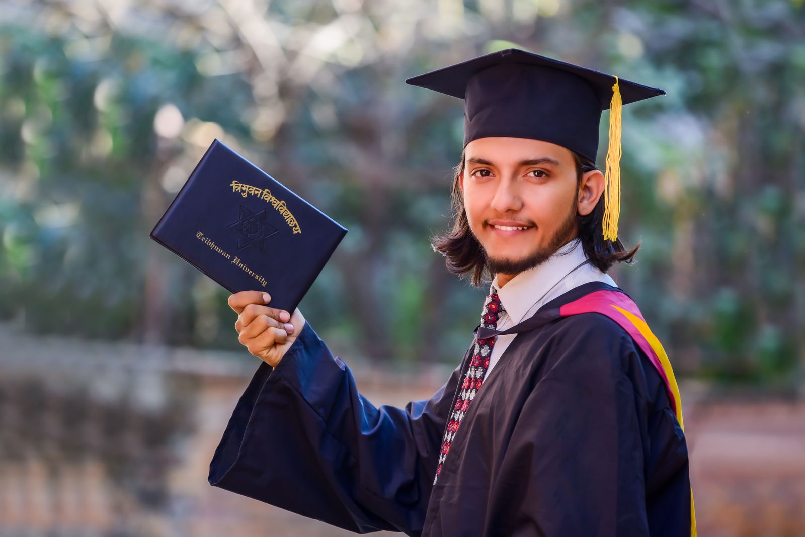A smiling graduate in a cap and gown holds up their diploma outdoors.