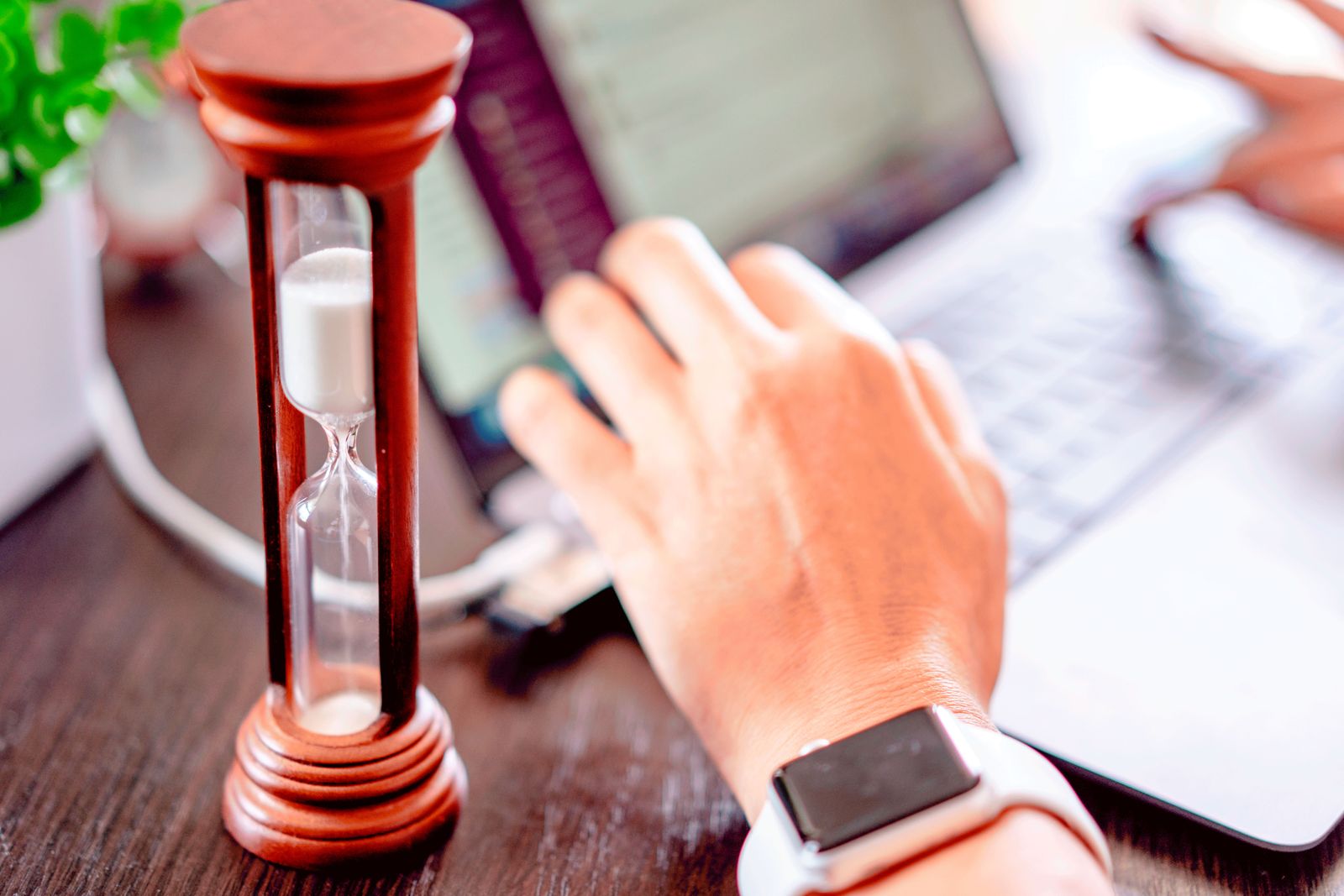 A close-up of a hand on a laptop keyboard next to an hourglass, symbolizing logged work hours.